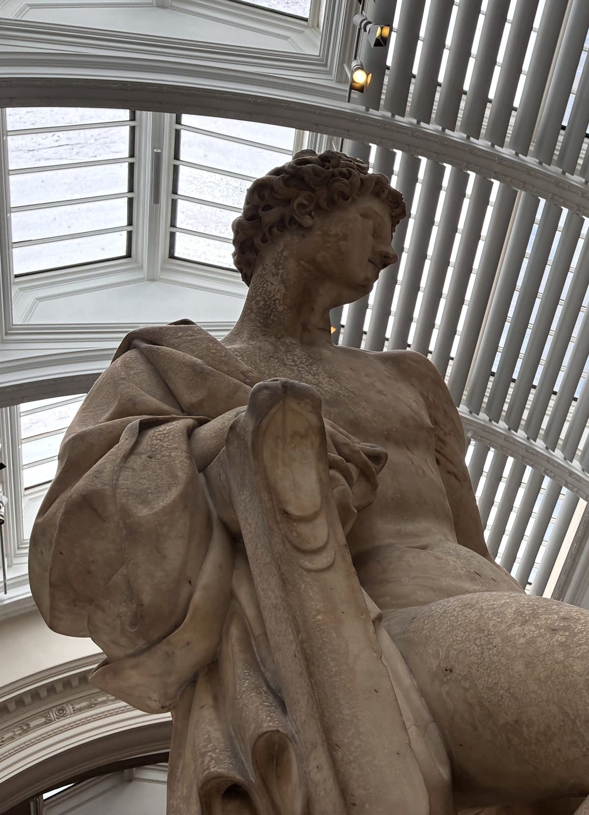 Marble statue of Apollo seen from below, robe over one shoulder, against the glazed roof of the V&A Renaissance gallery