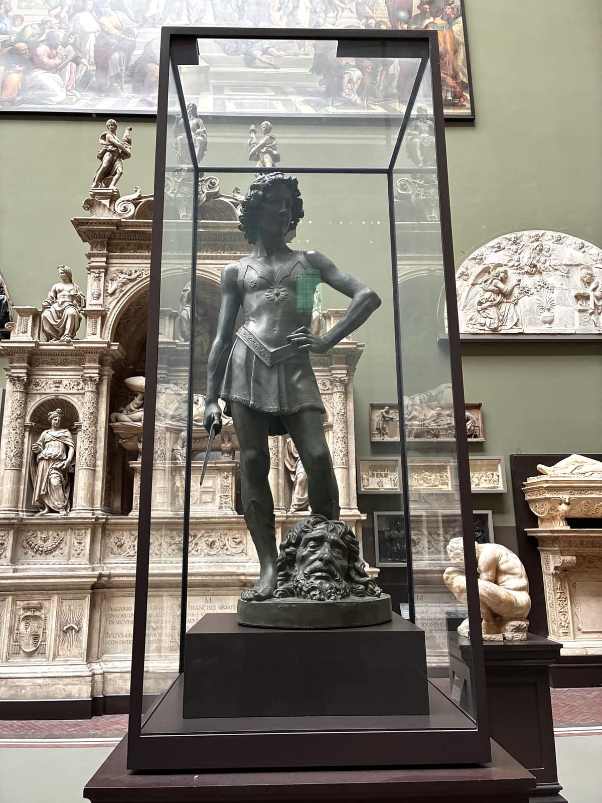 Cast of Verrocchio's David in a tall glass vitrine, wearing a short tunic with the head of Goliath at his feet