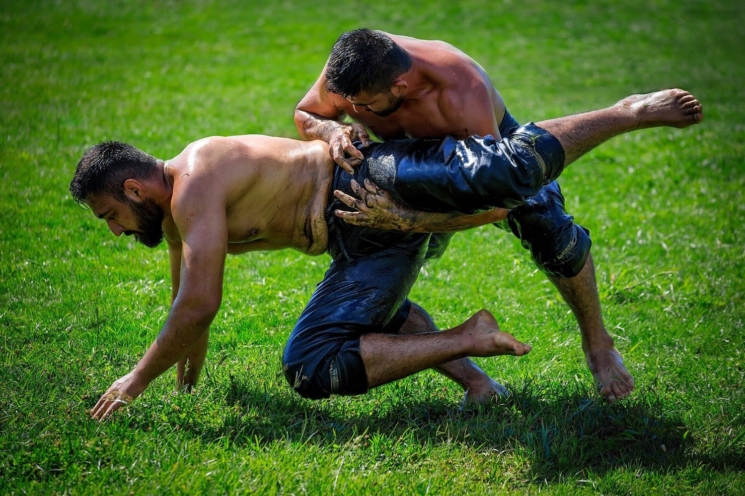 Two oiled Turkish wrestlers in leather kispet trousers grappling on grass, one sweeping the other off balance