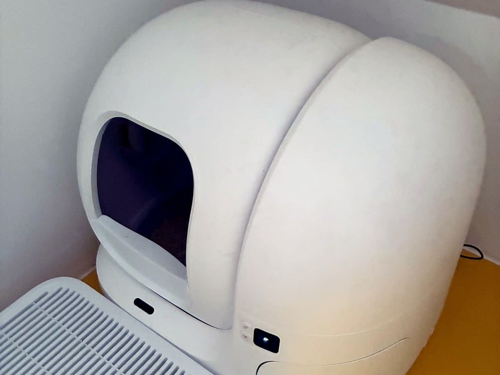 White PETKIT self-cleaning litter tray in the corner of a room beside an air purifier